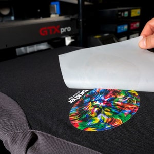 May include: A white sheet of transfer paper is being peeled off of a black t-shirt with a colorful graphic of a lion's head. The text "The roar of" and "Panther" are visible on the t-shirt.
