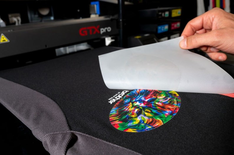 May include: A white sheet of transfer paper is being peeled off of a black t-shirt with a colorful graphic of a lion's head. The text "The roar of" and "Panther" are visible on the t-shirt.