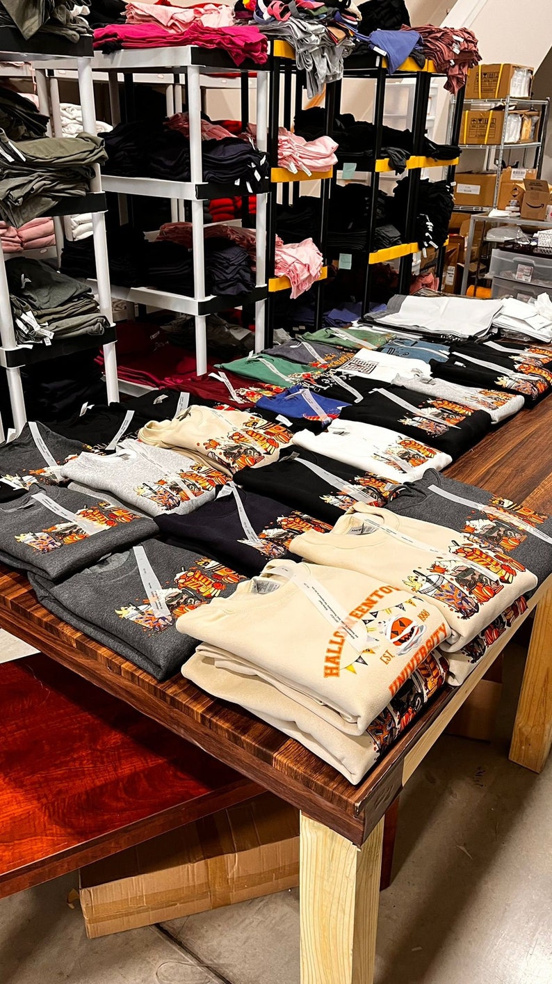 May include: A display of folded sweatshirts and t-shirts, featuring various colors and designs. Some shirts have a graphic print with text. The items are arranged on a wooden table and shelves, suggesting a retail or storage setting.