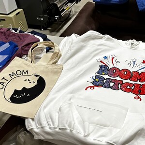 May include: Assortment of apparel including a white hoodie with "Boom Bitch" graphic, a blue t-shirt with a colorful design, a "Cat Mom" tote bag, and other t-shirts. The items are displayed on a table.