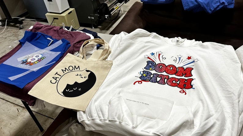 May include: Assortment of apparel including a white hoodie with "Boom Bitch" graphic, a blue t-shirt with a colorful design, a "Cat Mom" tote bag, and other t-shirts. The items are displayed on a table.