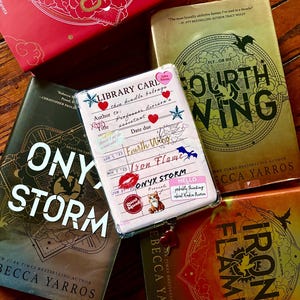 Puede incluir: Una colección de libros de Becca Yarros, incluyendo "Fourth Wing", "Onyx Storm" e "Iron Flame", dispuestos sobre una superficie de madera. Se observa una tarjeta de biblioteca con pegatinas. Los libros tienen cubiertas doradas, rojas y marrones con varios diseños y títulos.