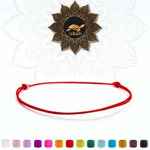 Könnte beinhalten: Ein minimalistisches rotes Schnurarmband mit goldenen Akzenten. Es wird vor einem weißen Hintergrund präsentiert, mit einem dekorativen schwarz-goldenen Logo. Das Logo zeigt eine Schildkröte und das Wort "jabuti".