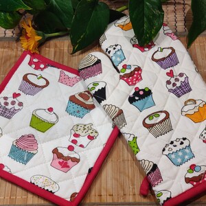 May include: Two white quilted oven gloves with red trim. Both oven gloves feature a pattern of colourful cupcakes with different designs.