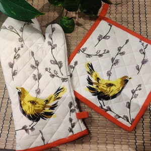 May include: A set of two white quilted oven mitts with orange trim. The mitts feature a floral pattern with yellow birds.