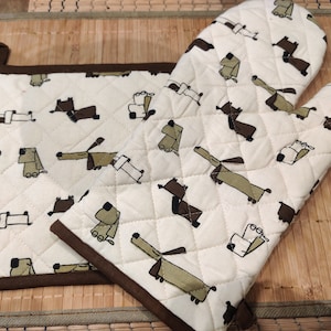 Könnte beinhalten: Zwei braune und weiße gesteppte Ofenhandschuhe mit einem Muster aus Comic-Hunden. Die Handschuhe sind aus Baumwolle und haben einen braunen Besatz.