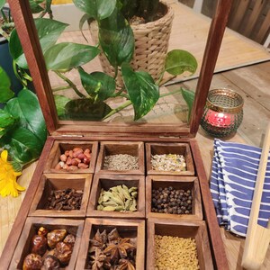 Peut inclure: Une boîte à épices en bois avec un couvercle en verre, divisée en 10 compartiments. Chaque compartiment contient une épice différente, notamment des cacahuètes, du cumin, des graines de fenouil, des gousses de cardamome, des grains de poivre noir, de l'anis étoilé et des graines de fenugrec.