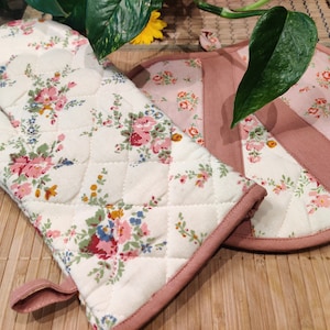 Può includere: Un set di due presine da cucina con stampa floreale. Una è in stile guanto e l'altra è un sottopentola rotondo. Entrambe sono trapuntate e hanno un bordo rosa.