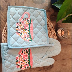 May include: A set of two blue quilted oven gloves with a floral teacup design. The oven gloves are made of cotton and have a quilted texture.