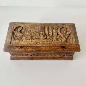 May include: A rectangular wooden box with detailed carvings on the lid. The box is a warm brown colour and features a relief of a bird and foliage. The box has a raised base and a slightly darker tone on the sides. The box is approximately 15 cm long.