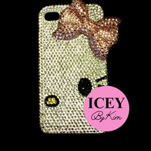 Puede incluir: Una funda de teléfono adornada con pedrería con un lazo y un diseño de cara de dibujos animados. La funda está cubierta de pequeños y brillantes pedrería. El lazo es una mezcla de pedrería dorada y rosa. El texto "ICEY By Kim" está en un círculo rosa.
