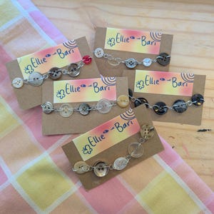 Puede incluir: Pulseras de botones exhibidas en tarjetas pequeñas. Cada pulsera presenta una serie de botones de varios colores y tamaños, conectados por una cadena plateada. Las tarjetas tienen un fondo degradado colorido con el texto "Ellie-Bari".