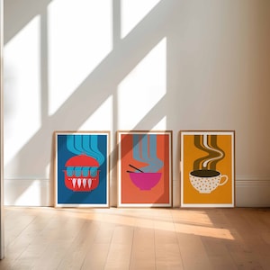May include: Three framed prints with a retro design. Each print features a different food item with steam rising from it. The first print shows a red pot with a lid on a blue background. The second print shows a pink bowl with chopsticks on an orange background. The third print shows a white cup with brown coffee on a yellow background.