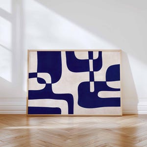 May include: Abstract art print featuring a geometric design in navy blue and off-white. The artwork is framed in a light wood frame and leans against a white wall. The design consists of curved shapes and lines.