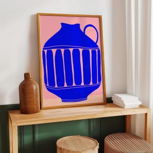 May include: A framed wall art print featuring a vibrant blue graphic illustration of a vase with vertical stripes and a single handle, set against a solid light pink background. The artwork is displayed in a thin wooden frame.