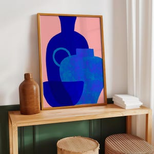 May include: A framed art print featuring a graphic illustration of three overlapping vessels in shades of deep blue and bright blue against a solid light pink background. The artwork is displayed in a thin, light-colored wooden frame.