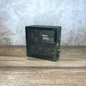 May include: A compact black computer case featuring a prominent hexagonal honeycomb mesh side panel. The rectangular chassis has a solid frame with visible screws at the corners and a side-mounted input and output port section with gold connectors.