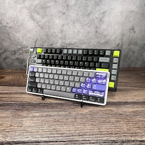 May include: Two mechanical keyboards on a wooden surface. The larger keyboard is black with lime green accents. The smaller keyboard is white with gray and purple keycaps. The keyboards are displayed against a gray textured background.