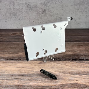 May include: A white metal computer component with multiple circular holes and the text "fractal design" printed on it. A small black component is below it. The component is on a wooden surface.