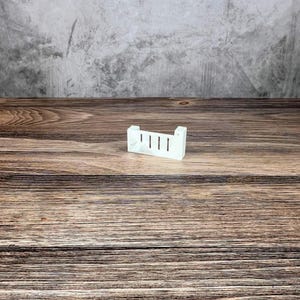 May include: A small, white, rectangular object with four vertical slots. The object has a simple, minimalist design and is placed on a wooden surface. The background is a textured grey wall.