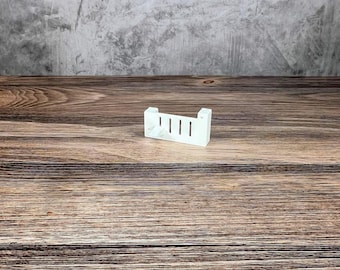 May include: A small, white, rectangular object with four vertical slots. The object has a simple, minimalist design and is placed on a wooden surface. The background is a textured gray wall.