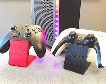 Könnte beinhalten: Ein schwarzer Gaming-Controller steht auf einem roten Ständer und ein weiß-schwarzer Gaming-Controller auf einem schwarzen Ständer. Beide Ständer tragen den eingeprägten Schriftzug The3DPC auf der Vorderseite. Dahinter steht ein PC-Gehäuse mit RGB-Licht.