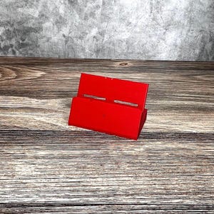 May include: A bright red, angled phone stand made of a solid material. The stand has two horizontal slots and is positioned on a wooden surface. The background features a textured gray wall.