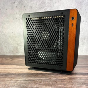 Peut inclure: Un boîtier d'ordinateur noir avec un panneau avant en maille nid d'abeille et un panneau latéral en bois. Le boîtier possède un bouton d'alimentation et un port USB. Il est posé sur une surface en bois.