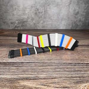 Op de afbeelding: Twee sets computerkabelverlengingen met kleurrijke kabelkammen. De bovenste set heeft witte kabels met roze, grijze, rode, gele, zwarte, witte, blauwe en oranje kammen. De onderste set heeft zwarte kabels met oranje, blauwe, witte en gele kammen.