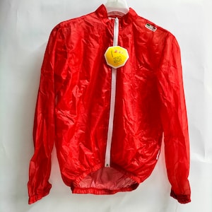 Op de afbeelding: Rode lichtgewicht fietsjas met een witte rits en een geel label met de tekst "Zero Wind".