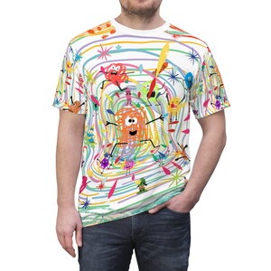 May include: A white t-shirt featuring a colourful, childlike design with a variety of cartoon characters and abstract shapes. The design includes a central orange character with a large smile and eyes, surrounded by other colourful characters and lines.