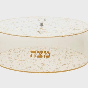 May include: A clear, round cake stand with a lid. The stand has a gold-colored rim and a gold speckled pattern on the base and lid. The word "Matzah" is printed in gold on the base. A crystal knob is on top of the lid.