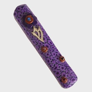 May include: A purple mezuzah case with a floral design and a silver-colored emblem. The case has a repeating pattern of dark purple shapes and is adorned with three brown and red accents.