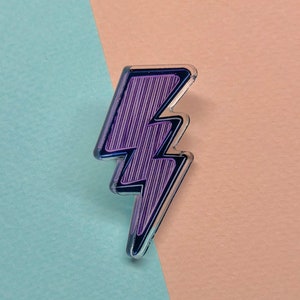 Peut inclure: Une épingle en forme d'éclair violet et noir. L'épingle a une bordure transparente et un motif rayé à l'intérieur de l'éclair. Le fond est divisé en bleu clair et pêche.