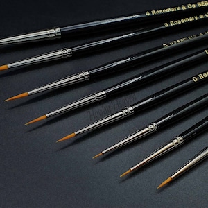 May include: A set of six black handled paint brushes with gold lettering. The brushes are labelled 0, 1, 3, and 4 Rosemary & Co.  The brushes are arranged in a row on a black surface.