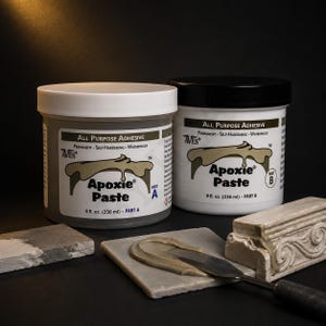 May include: Two 236 ml containers of Aves Apoxie Paste, labelled Part A and Part B, featuring white and black lids. The product is an all-purpose adhesive. A metal spatula rests on a square tile with a smear of the paste next to a decorative moulding.