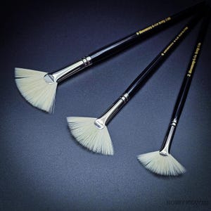Op de afbeelding: Drie waaiervormige penselen met witte haren, zilverkleurige metalen hulzen en zwarte houten stelen. Op elke steel staat in goudkleurige letters Rosemary & Co Ivory Fan. De penselen variëren in grootte en zijn diagonaal gerangschikt.