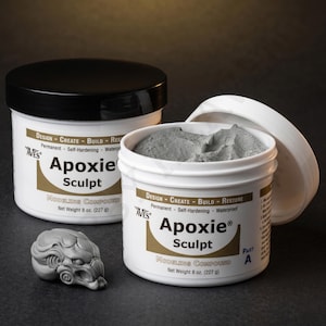 May include: Two white tubs of Apoxie Sculpt modelling compound, one with a black lid and the other open, showing grey compound. A grey sculpted figure is in front. The tubs have text that reads "DESIGN - CREATE - BUILD - RESTORE". Each tub contains 227g.