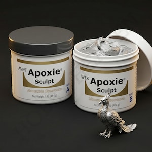 May include: Two white containers of Aves Apoxie Sculpt modeling compound, one with a closed lid and the other open, revealing gray material. A silver phoenix sculpture sits in front. Each container is labeled with "Net Weight 1 lb (454 g)".