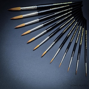 Może przedstawiać: Zestaw 14 pędzli z czarnymi uchwytami i złotym napisem. Pędzle są ułożone wachlarzowo na ciemnoszarym tle. Napis na uchwytach brzmi "Rosemary & Co Designer Red Dot".