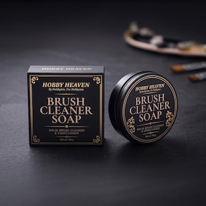 Op de afbeelding: Een zwarte vierkante doos en een rond zwart blik met Hobby Heaven penseelreinigingszeep. Beide zijn voorzien van gouden tekst met Hobby Heaven, By Hobbyists, For Hobbyists, Brush Cleaner Soap en Solid Brush Cleaner & Conditioner, met een nettogewicht van 100g.