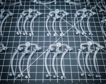 May include: A collection of clear, plastic, curved components arranged on a black grid surface. The components have a rounded top and a curved stem with small circular details at the base. The grid has numbered markings.