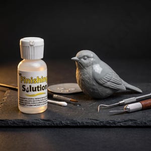 Op de afbeelding: Een grijs vogelbeeldje naast een fles "Finishing Solution" en diverse gereedschappen. De fles heeft een witte dop en een geel en wit etiket. Er is ook een klein, rond palet met vloeistof aanwezig.