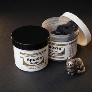 May include: Two white tubs of Apoxie Sculpt modeling compound with black lids and a silver cat figurine. One tub is open, revealing dark gray compound. The text on the tubs reads "Design, Create, Build, Restore".