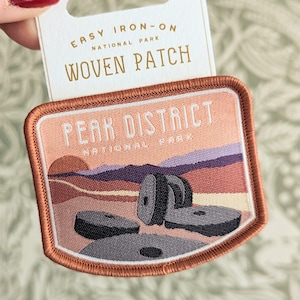 May include: A woven iron-on patch featuring a sunset scene with the text "Peak District National Park". The patch is orange, pink, and white with a brown border.