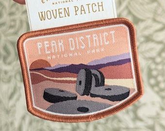 May include: A woven iron-on patch featuring a sunset scene with the text "Peak District National Park". The patch is orange, pink, and white with a brown border.