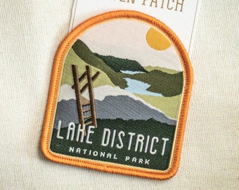May include: An orange and green woven patch with the text "Easy Iron-On National Park Woven Patch" and "Lake District National Park". The patch features a mountain scene with a ladder and a lake.