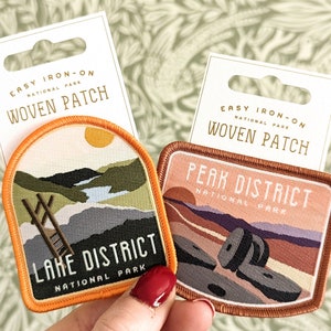 K&ouml;nnte beinhalten: Zwei B&uuml;gelbilder aus gewebtem Stoff mit Motiven von Nationalparks. Das erste Patch ist orange und zeigt eine Landschaft mit einer Leiter und dem Text "Lake District National Park". Das zweite Patch ist braun und zeigt eine Landschaft mit Felsen und dem Text "Peak District National Park".