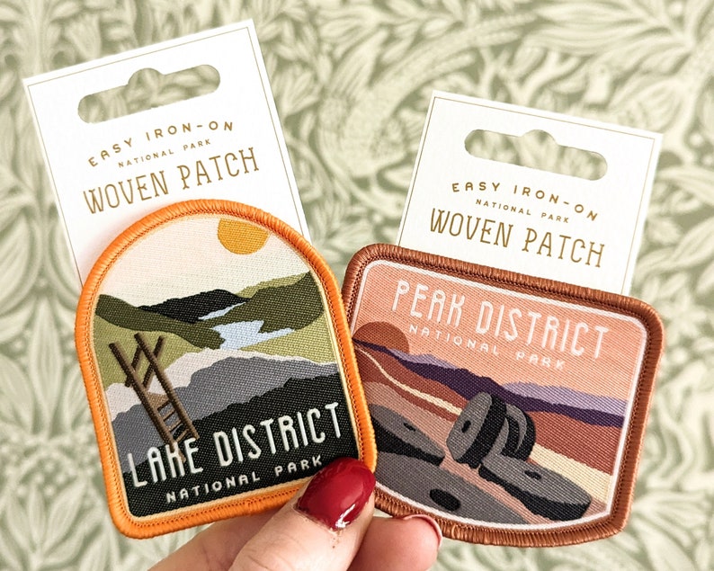 K&ouml;nnte beinhalten: Zwei B&uuml;gelbilder aus gewebtem Stoff mit Motiven von Nationalparks. Das erste Patch ist orange und zeigt eine Landschaft mit einer Leiter und dem Text "Lake District National Park". Das zweite Patch ist braun und zeigt eine Landschaft mit Felsen und dem Text "Peak District National Park".