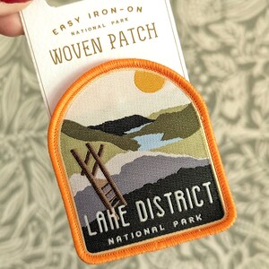 K&ouml;nnte beinhalten: Ein orangefarbener B&uuml;gel-Patch mit einer Landschaftszene eines Sees, Bergen und einer Leiter. Der Text "Lake District National Park" befindet sich auf dem Patch.